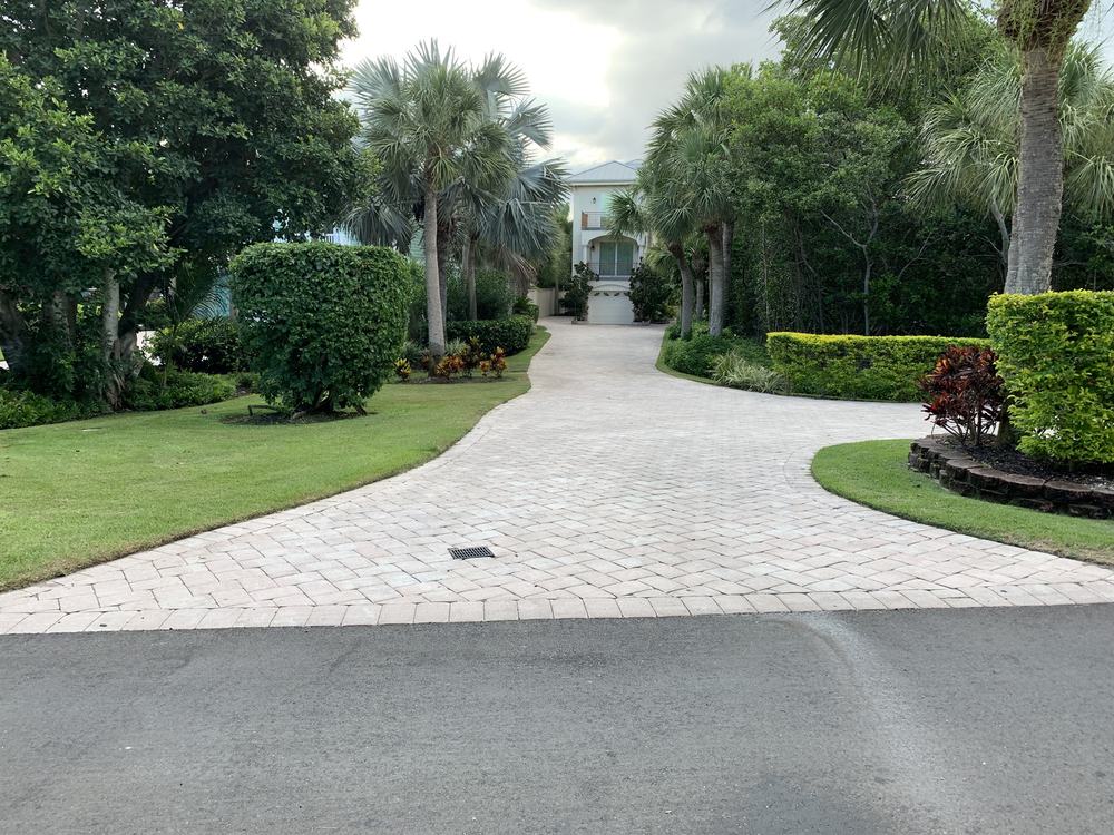 Paver driveway running between clipped hedges and palms to a screened entry